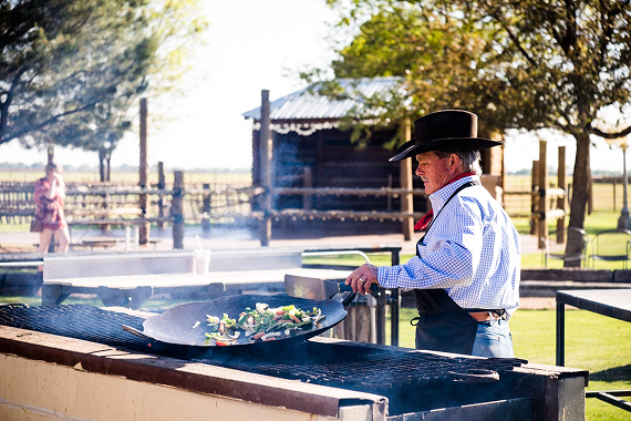 Chef cooking outdoors by a wooden cabin under sunlight.